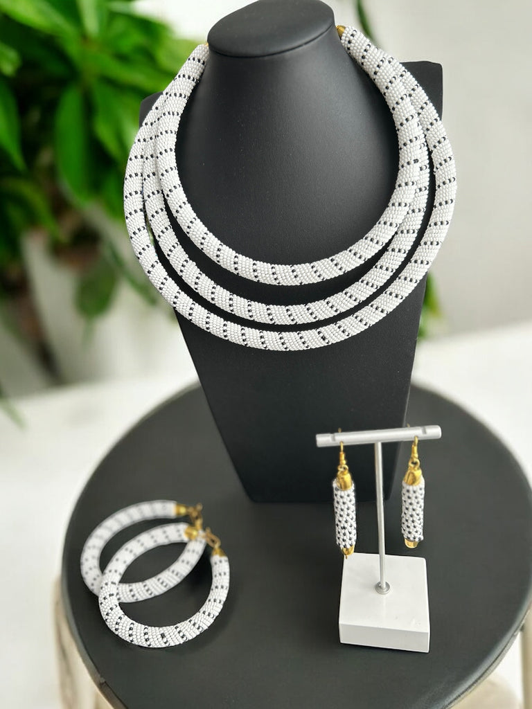 A white and black striped Maasai Regal necklace displayed on a mannequin bust, with a set of matching bracelets and earrings in gold tone.